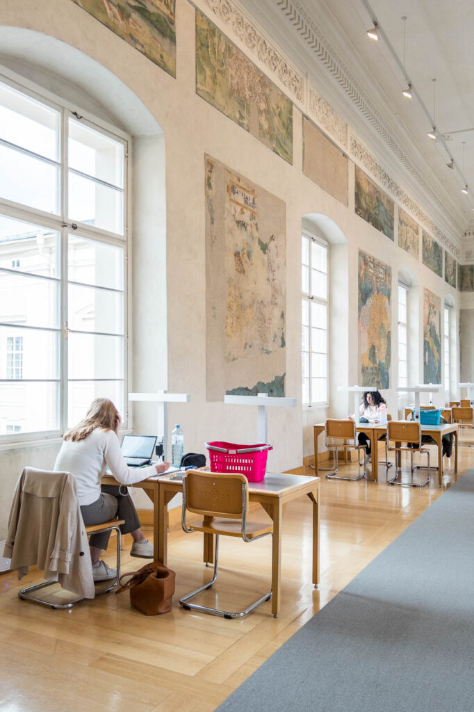 Interior of a library inside a Renaissance palazzo, showing a long and narrow reading room with work tables lining both walls. Between large vaulted windows, the walls are decorated with frescoes depicting maps.
