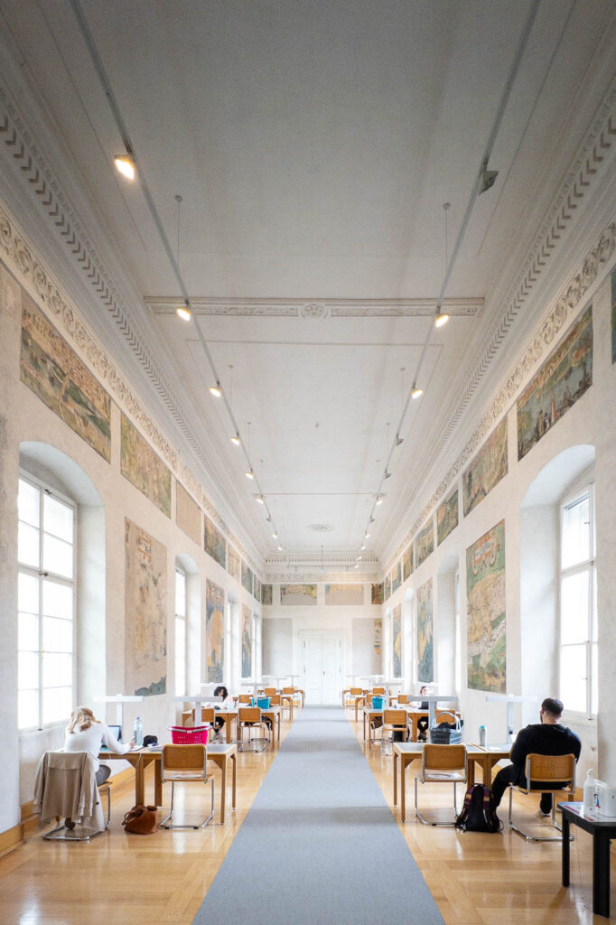 Interior of a library inside a Renaissance palazzo, showing a long and narrow reading room with work tables lining both walls. Between large vaulted windows, the walls are decorated with frescoes depicting maps.