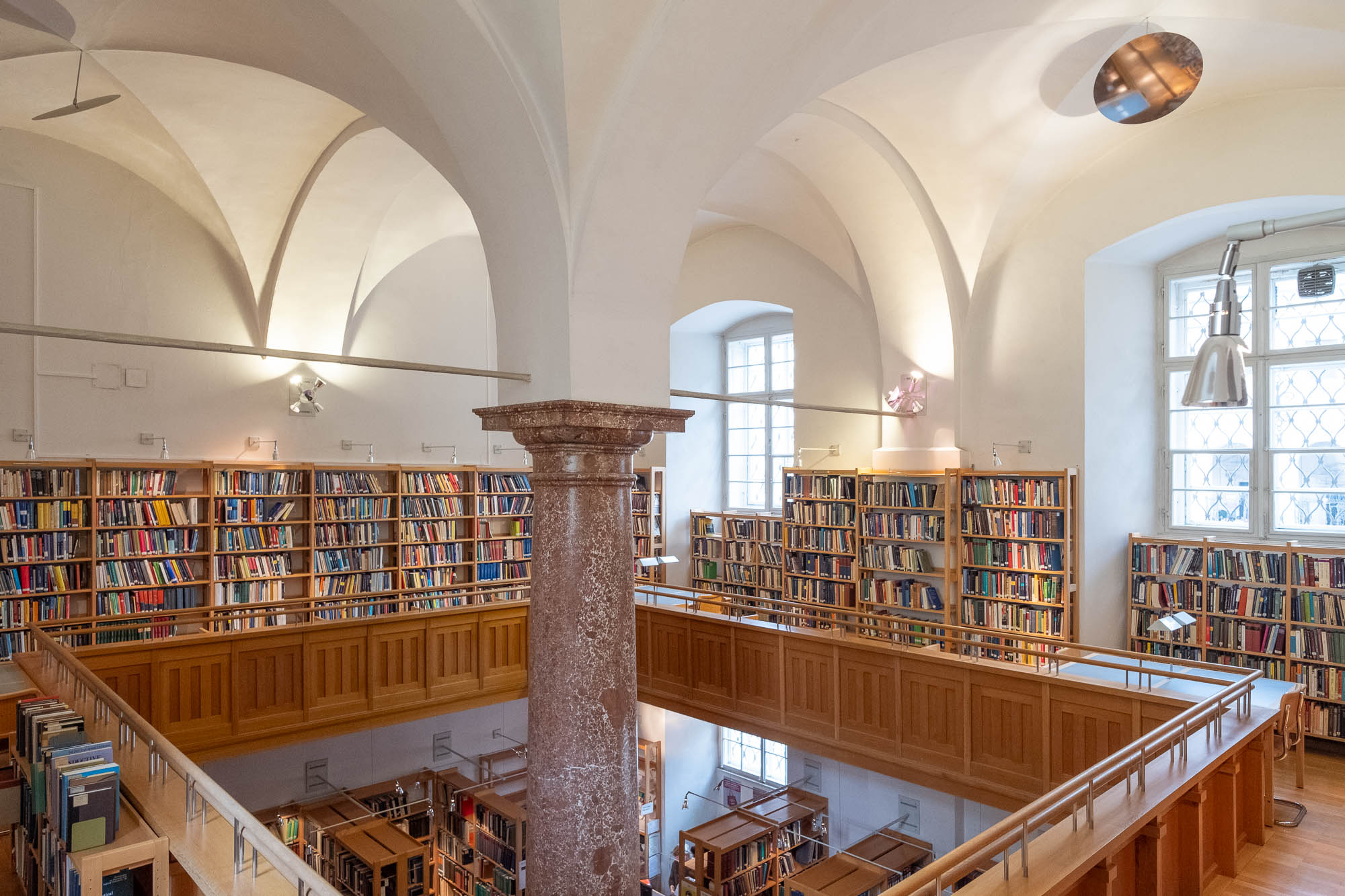 Interior of a library inside a Renaissance palazzo. The room has vaulted ceilings supported by a single large red marble column and a mezzanine with bookshelves and work tables around it.