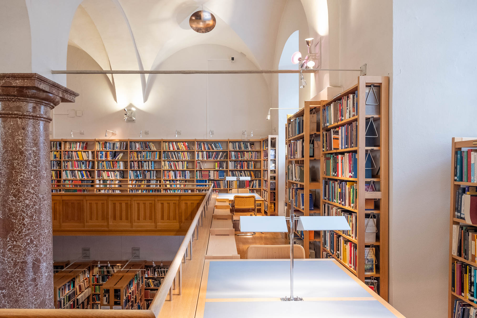 Interior of a library inside a Renaissance palazzo. The room has vaulted ceilings supported by a single large red marble column and a mezzanine with bookshelves and work tables around it.