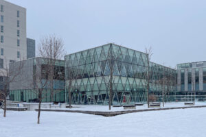 Exterior of a contemporary glass building, a two-storey structure with glazing supported by zigzag trusses. The sky is overcast and the building is surrounded by snow.