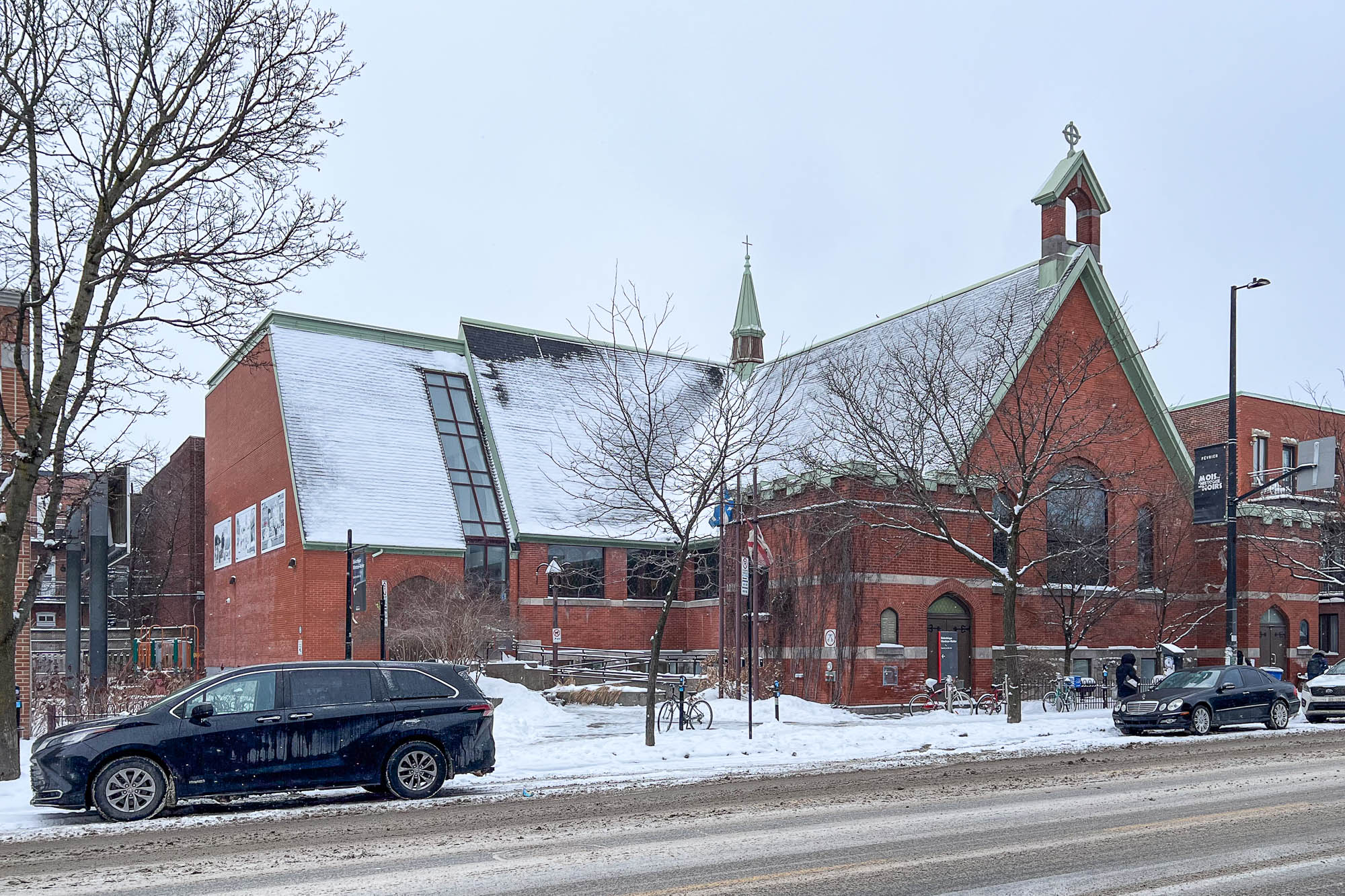Exterior of a brick church with a side extension, on a snowy street with cars parked in the front.