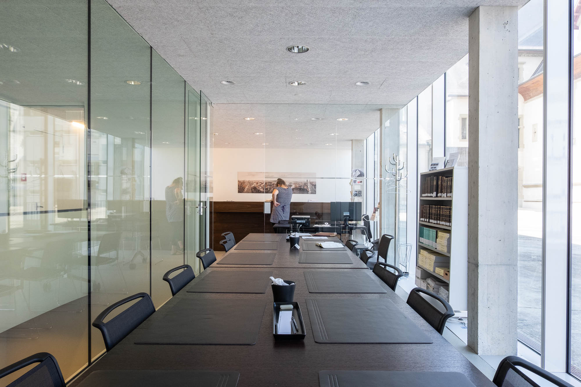 Interior of an archives reading room in a contemporary builindg, a long table with a glass partition to the left and large windows to the right. A person is standing at the back.