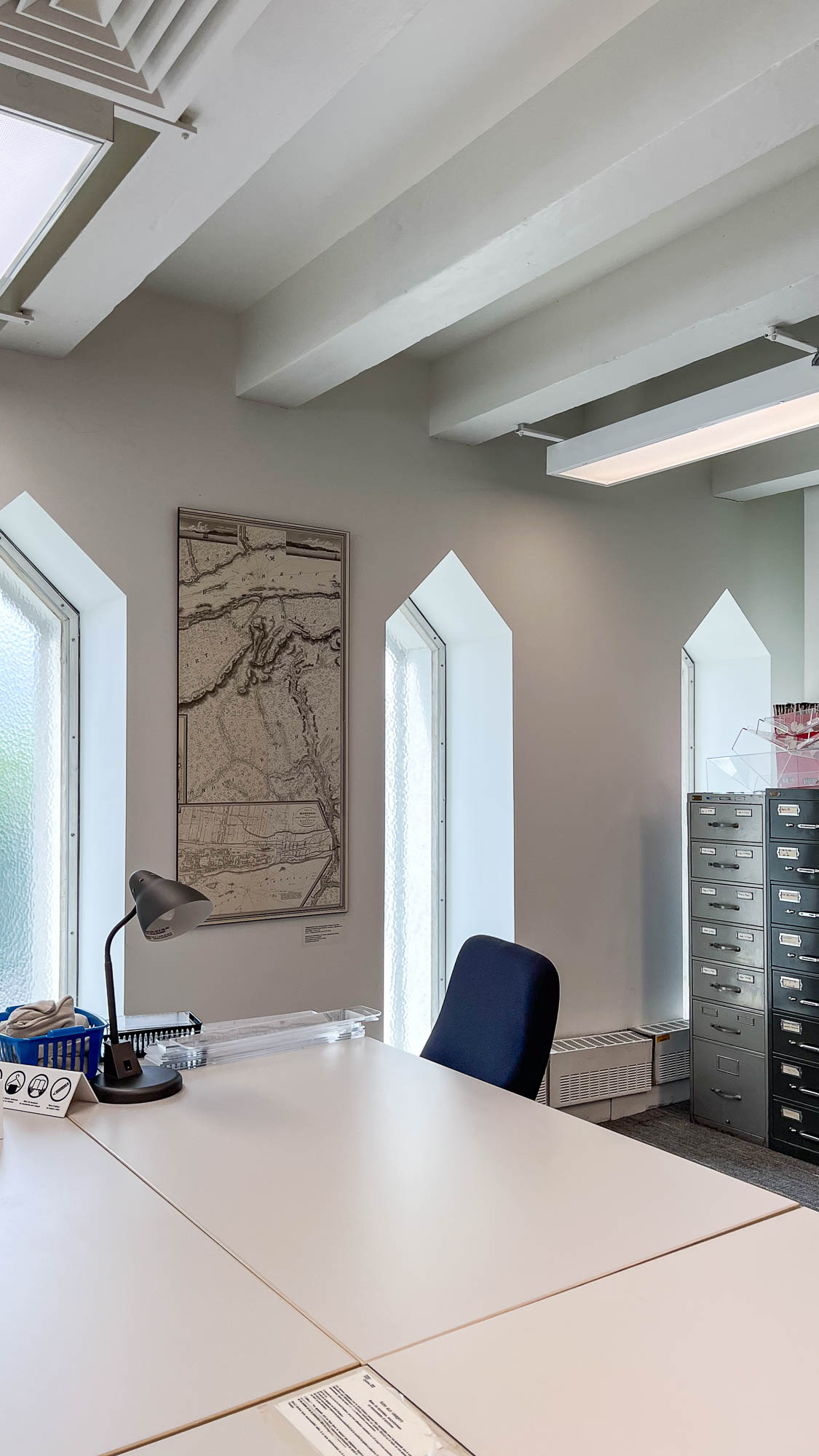 Interior of an archives reading room, a large table in the forefront with metal cabinets at the back. The space is lit by narrow pointed windows.
