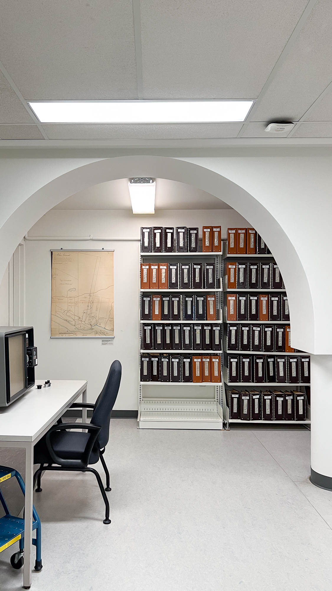 Interior of an archives reading room. Metal bookshelves bearing archival boxes face the back wall behind an arched doorway. A table with a microfiche reader is on the left.