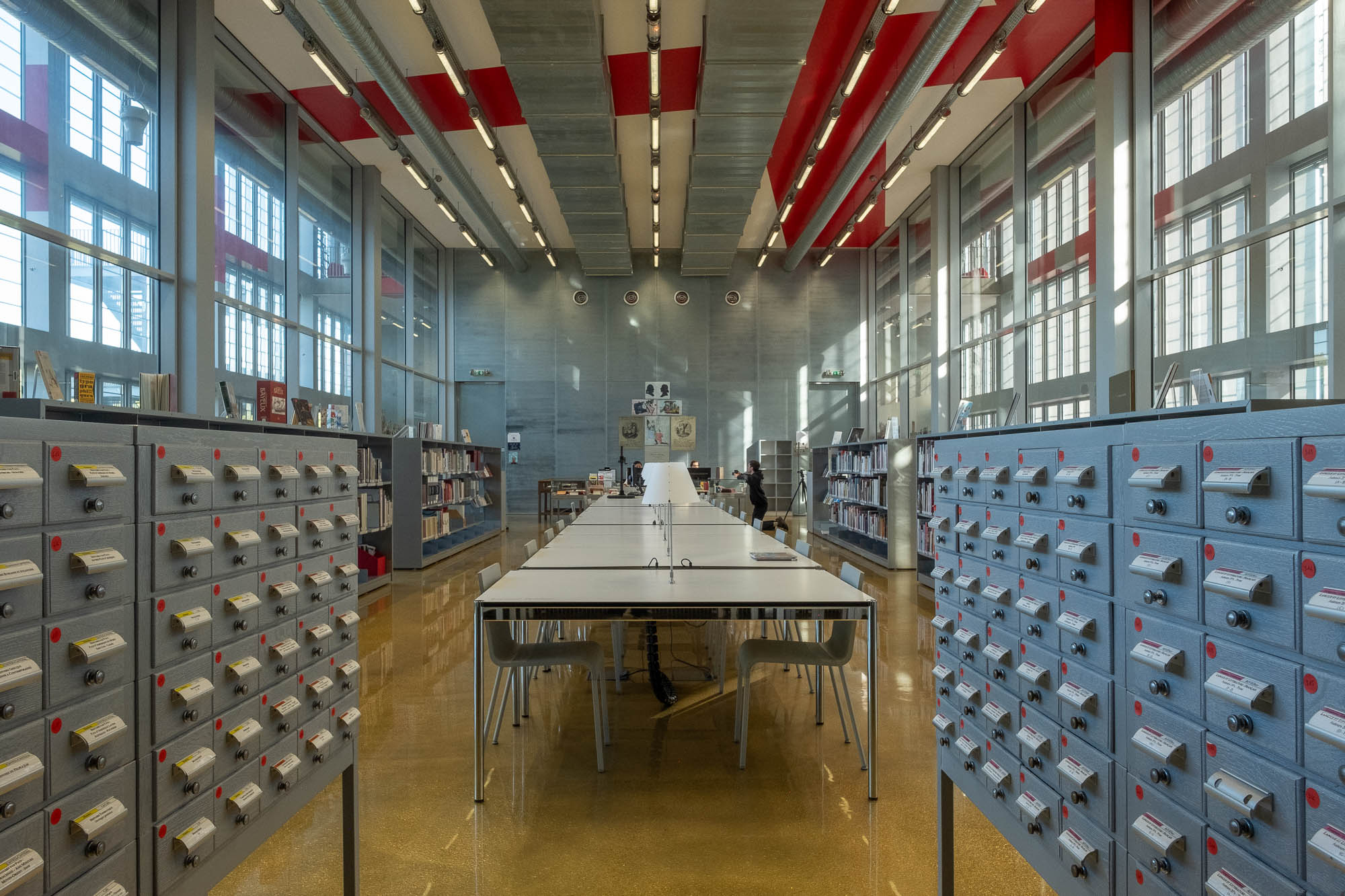 Interior of an archives reading room, a glass partitioned room in an industrial building with large windows on both sides. The room is filled with a row of tables in the middle, and shelves on the side. At the forefront are two card catalog cabinets.