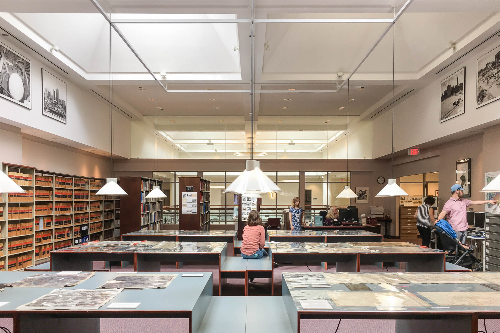 Interior of an archives reading room during a doors open event. Maps and documents are displayed on tables around which there are no chairs. Triangular glass lamps hang above the tables. Visitors are seen in the background looking at exhibits, a person is seated on a table in the middle of the image.