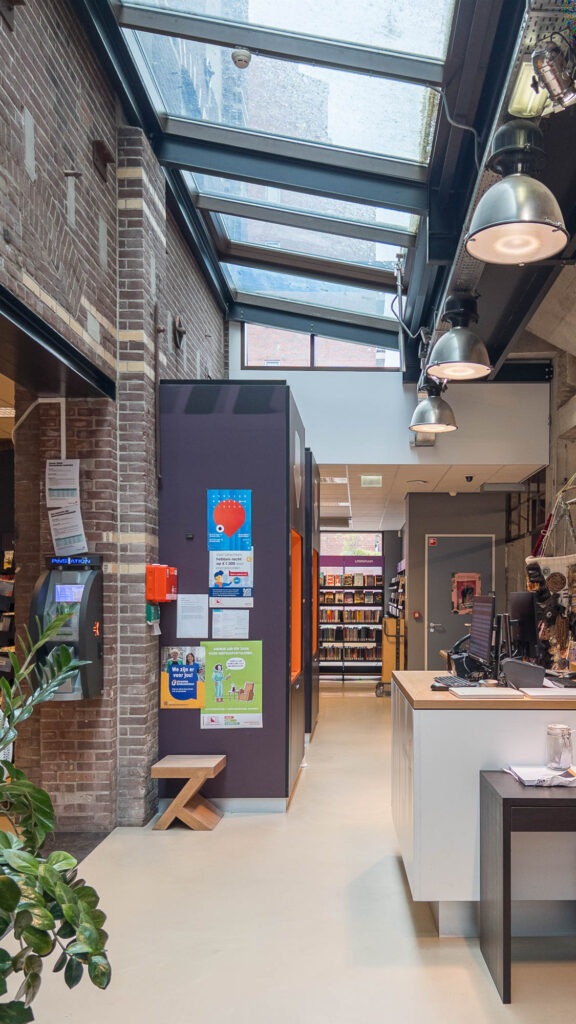Interior of a library in a converted industrial building with a glass ceiling and exposed brick walls. A reception desk is on the right, and bookshelves are in the background.