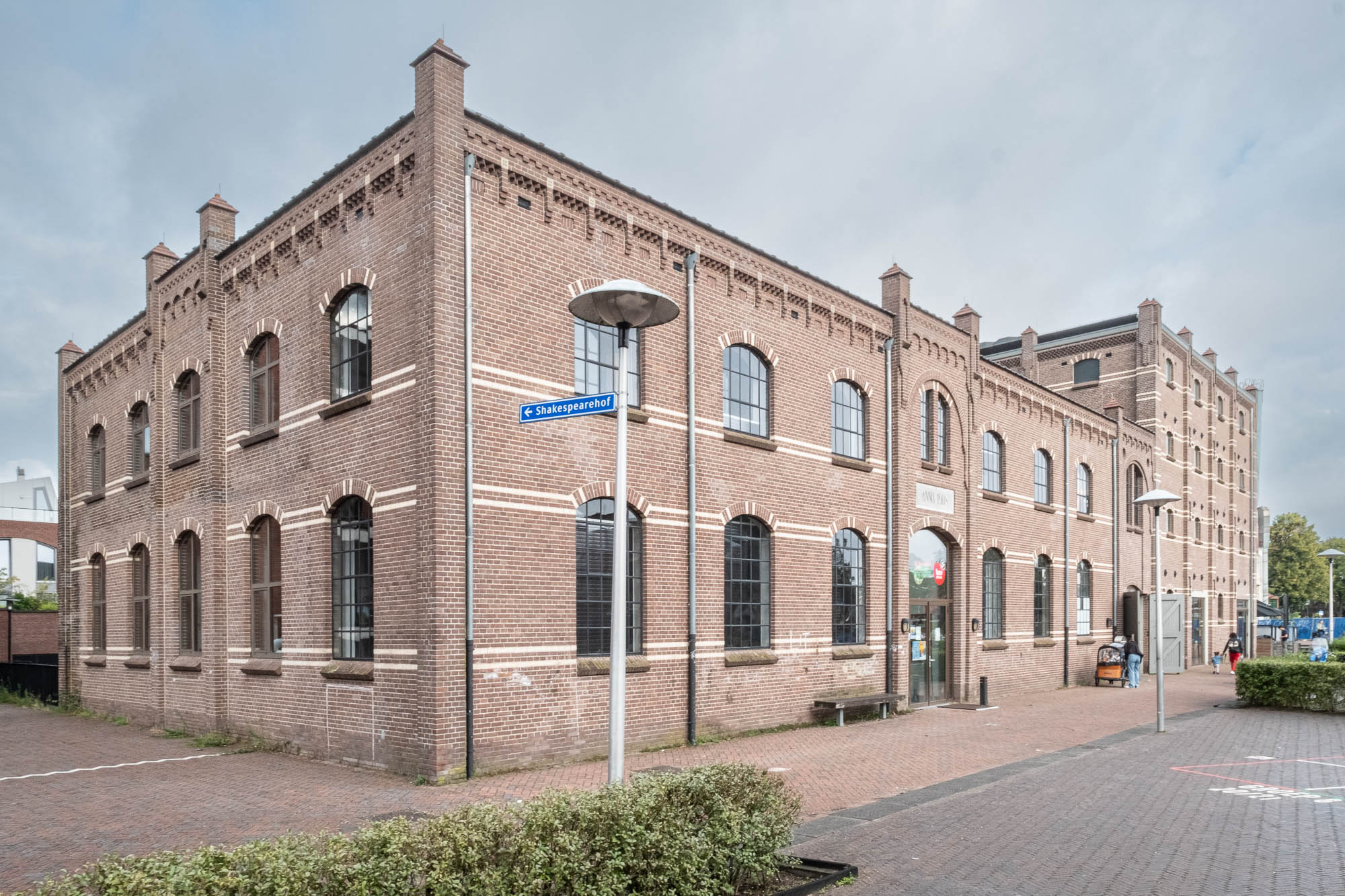 A large, historic brick building with arched windows and decorative brickwork stands under a cloudy sky. A street lamp and street sign reading "Shakespearehof" are in the foreground, and bushes line the sidewalk.