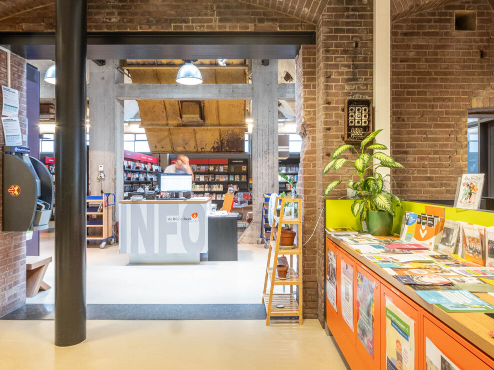 Interior of a library in a converted industrial building with exposed brick walls. A central information desk is staffed by a person. Shelves filled with books surround the area. A display with pamphlets is on the right.