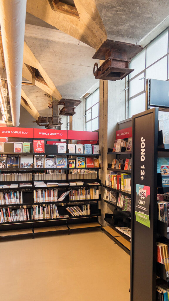 A library section in a converted industrial building with shelves filled with books. Signs indicate areas for different age groups, including Jong 12-, and a Werk & Vrije Tijd section.