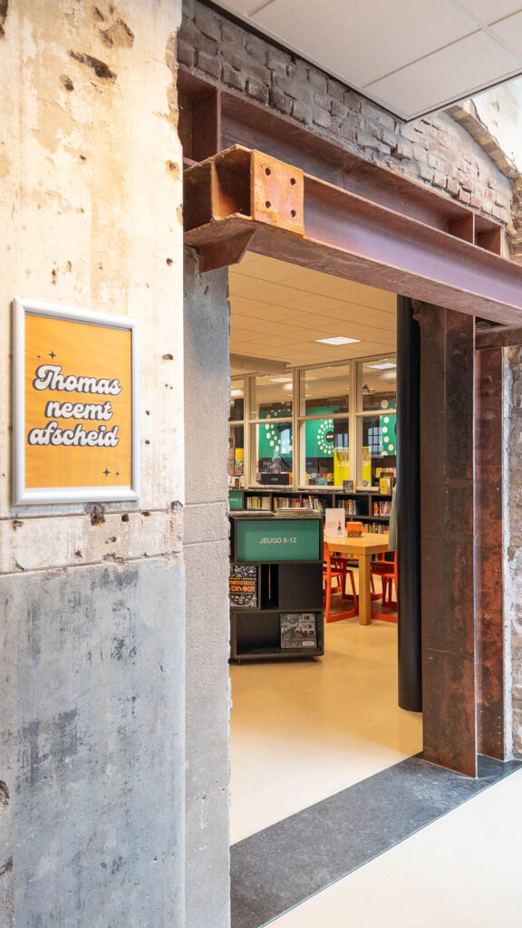 Entrance to a library in a converted industrial building with a brick and concrete wall. A poster on the left reads Thomas neemt afscheid. Inside, there are bookshelves, a sign JEUGD 9-12, and a few tables with red chairs. The space is well-lit with overhead lighting.