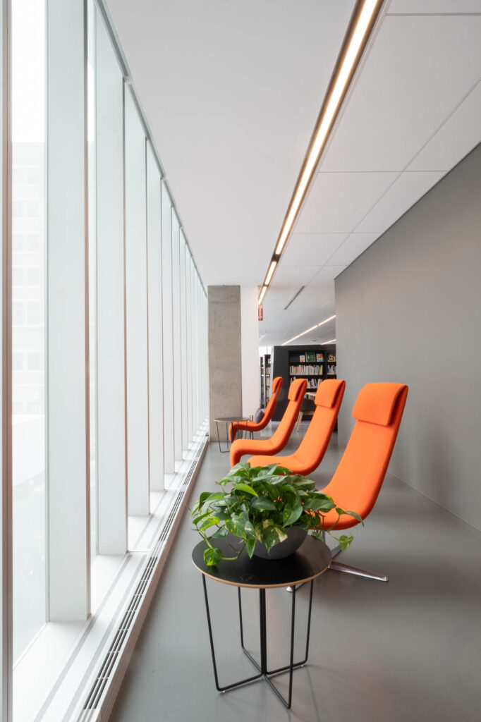 A modern interior features a row of orange lounge chairs beside a large window. A round table with a green potted plant is in the foreground.