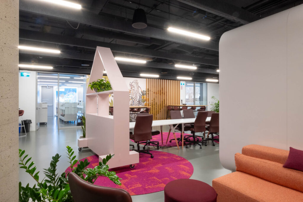 An office space with modern furnishings, featuring a white desk area with multiple chairs and a shelving unit resembling a house with a triangular roof. There are potted plants, colourful rugs, and a cozy seating area in the front.