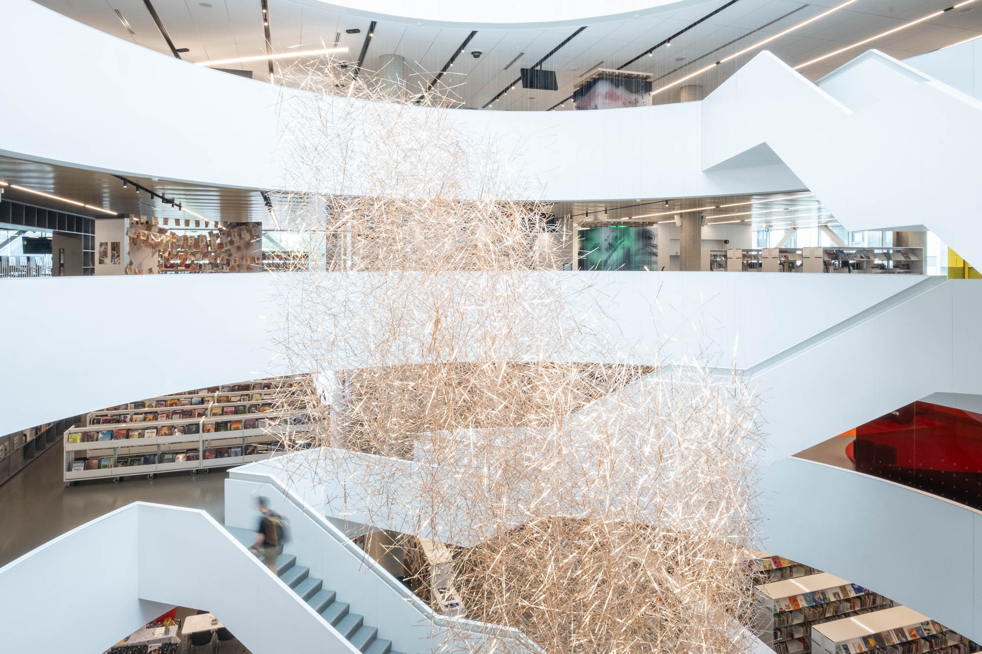 A modern library interior with several open floors connected by white staircases in a large circular atrium. Bookshelves line the walls on each level. An intricate art installation made of thin strips of brass is suspended in the central open space. A person is visible on the stairs.