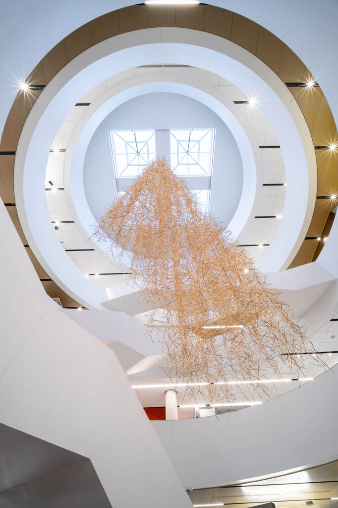 View from below of a modern interior with a circular skylight and large geometric sculpture made of thin strips of brass hanging from the ceiling.