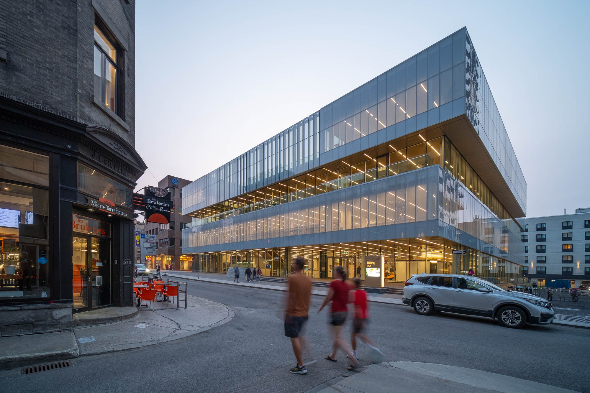 A modern glass and steel building with multiple stories stands on a street. People walk across the street, and cars are parked nearby. To the left is a café in a brick building with outdoor seating.