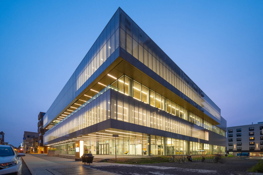 A modern, angular glass and steel building is lit from within at dusk, with large, illuminated windows and straight lines. The surrounding area includes a sidewalk, bicycles, and other buildings in the background.