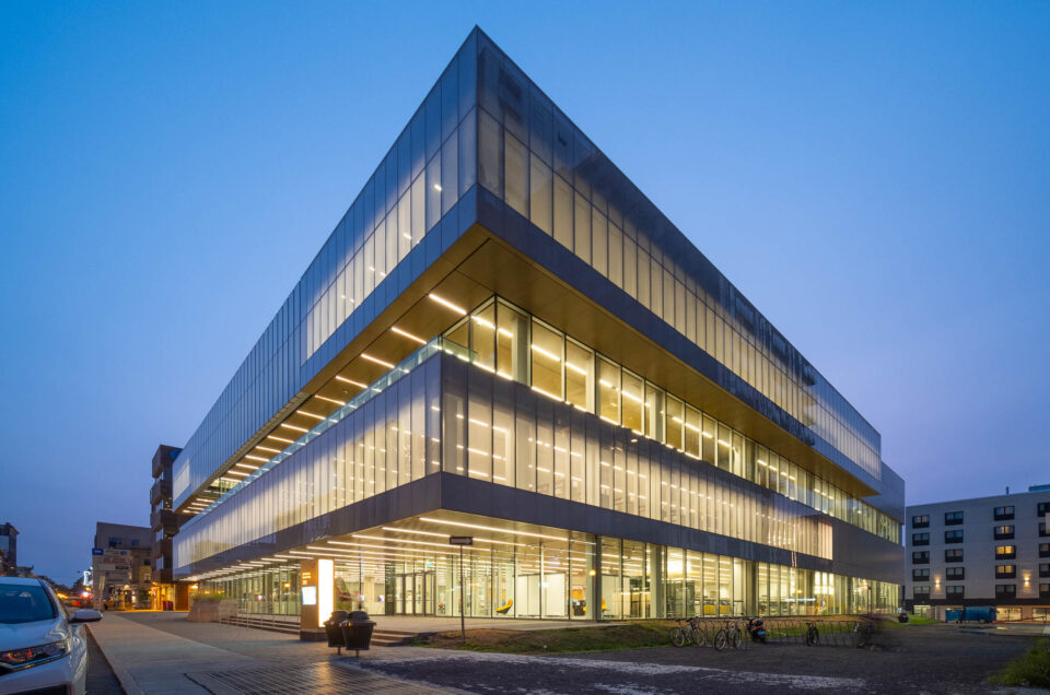 A modern, angular glass and steel building is lit from within at dusk, with large, illuminated windows and straight lines. The surrounding area includes a sidewalk, bicycles, and other buildings in the background.