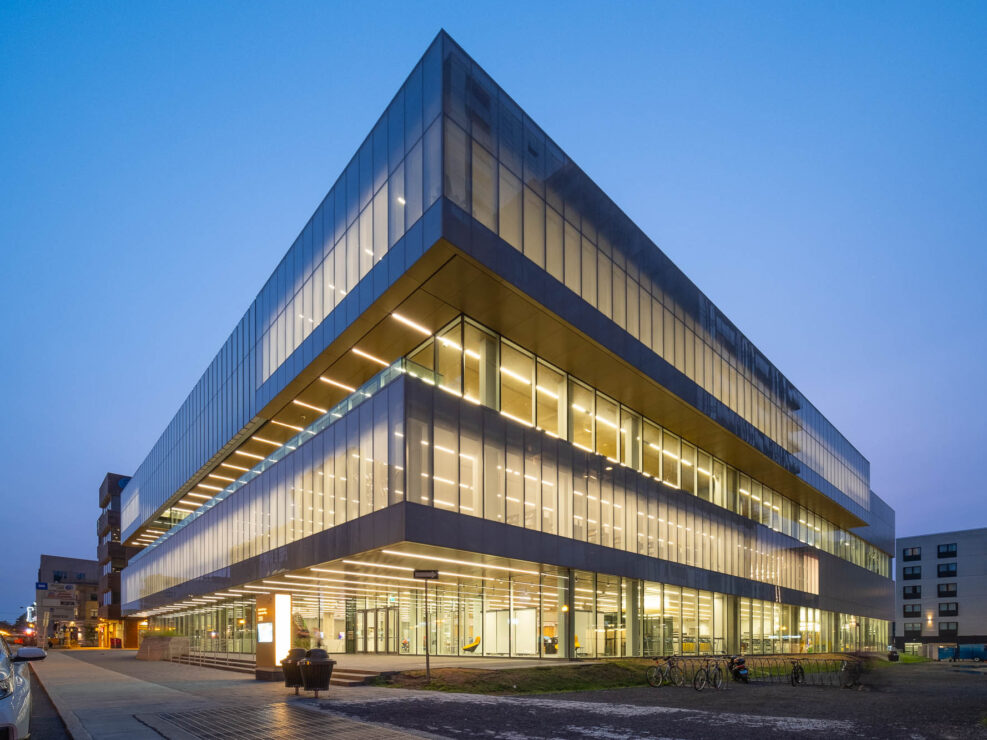 A modern, angular glass and steel building is lit from within at dusk, with large, illuminated windows and straight lines. The surrounding area includes a sidewalk, bicycles, and other buildings in the background.