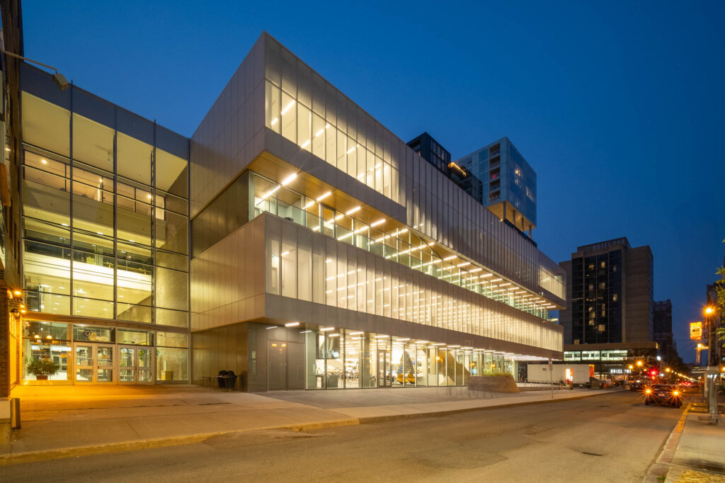 A modern, multi-story building with large glass windows is illuminated at dusk. The structure features angular architectural design elements. Cars and street signs are visible on the adjacent road, and city buildings are in the background.