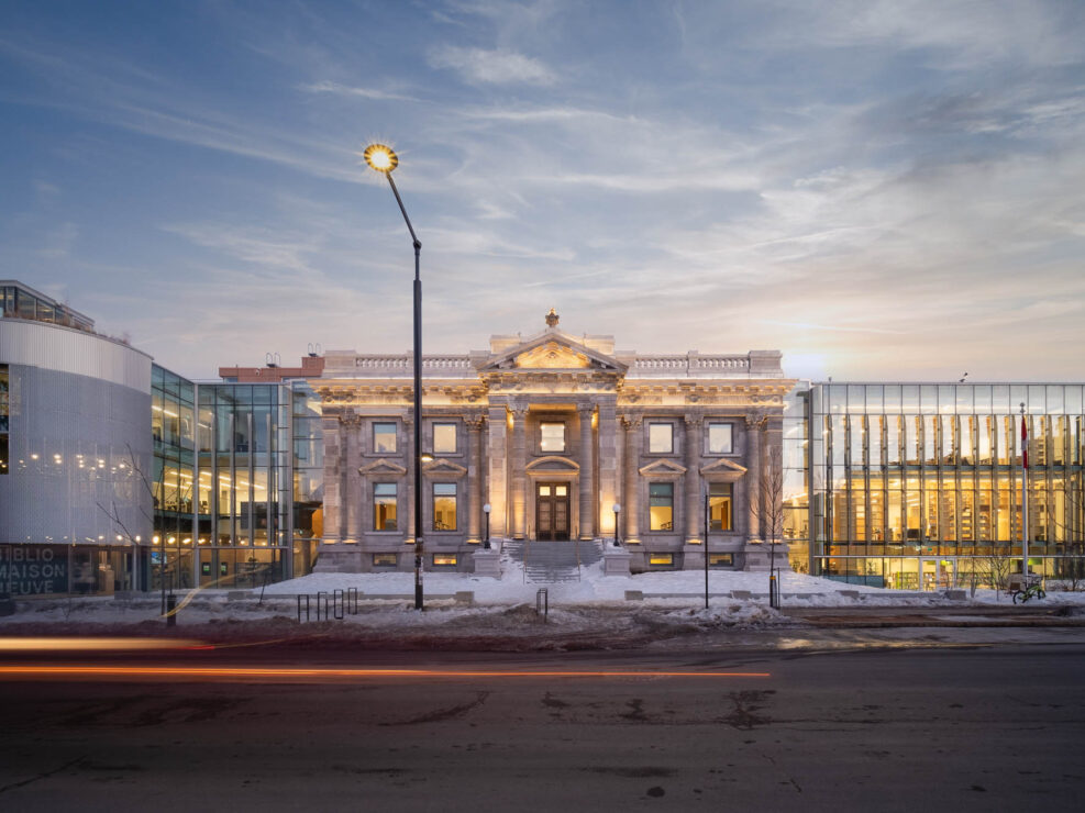 A historic building with classical architecture is flanked by modern glass structures on both sides. Snow covers the ground, and a blurred car passes by on the road in front. The scene is illuminated by soft evening light.