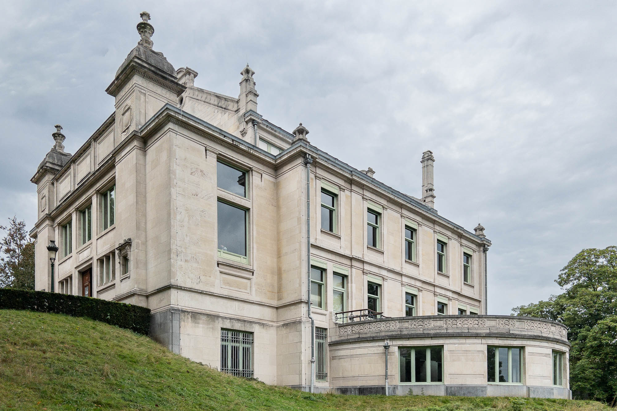A Neoclassical stone building on sloped terrain with twin turrets crowned with decorative urns. A semi-circular extension topped by a terrace extends from the lower level. The sky is overcast.