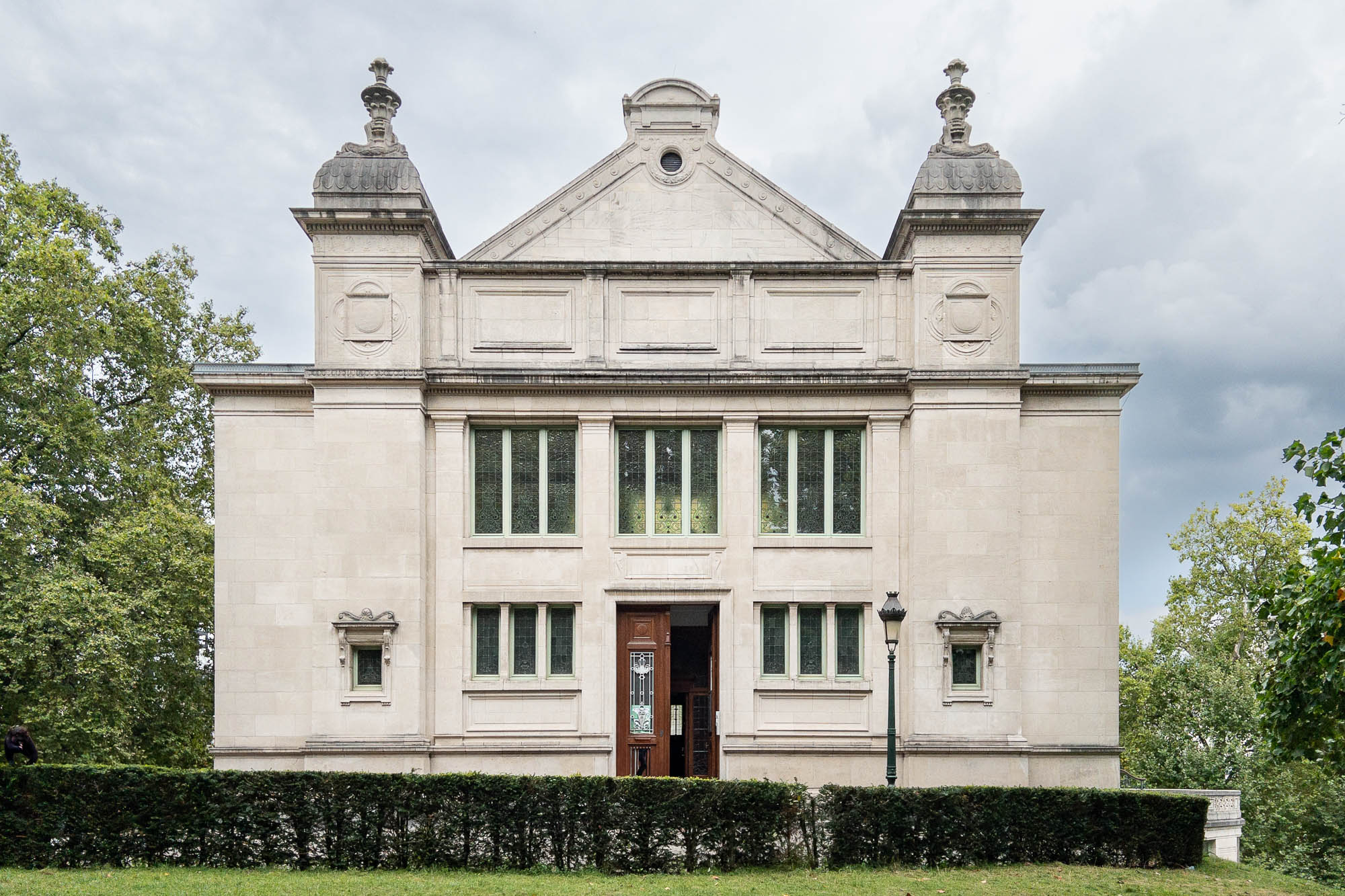 A symmetrical stone building with classical architectural features with a triangular roof line flanked by two turrets topped with decorative urns. It has tall windows and a wooden door and is surrounded by trees and a neatly trimmed hedge in the foreground.