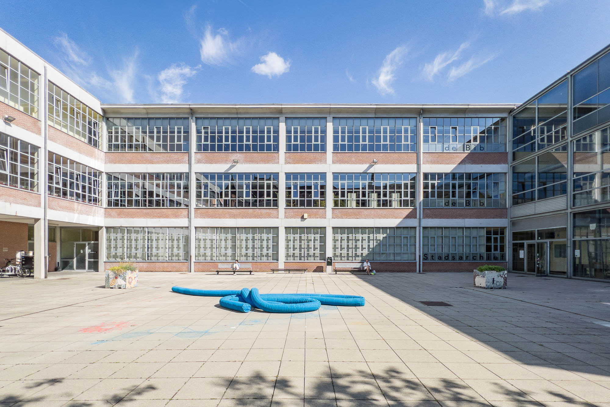A courtyard with a large, three-story industrial-style building in the background with a facade of brick and glazing. The courtyard is paved with concrete tiles, and a blue, tube-like structure is placed in the centre. The sky is clear and sunny.