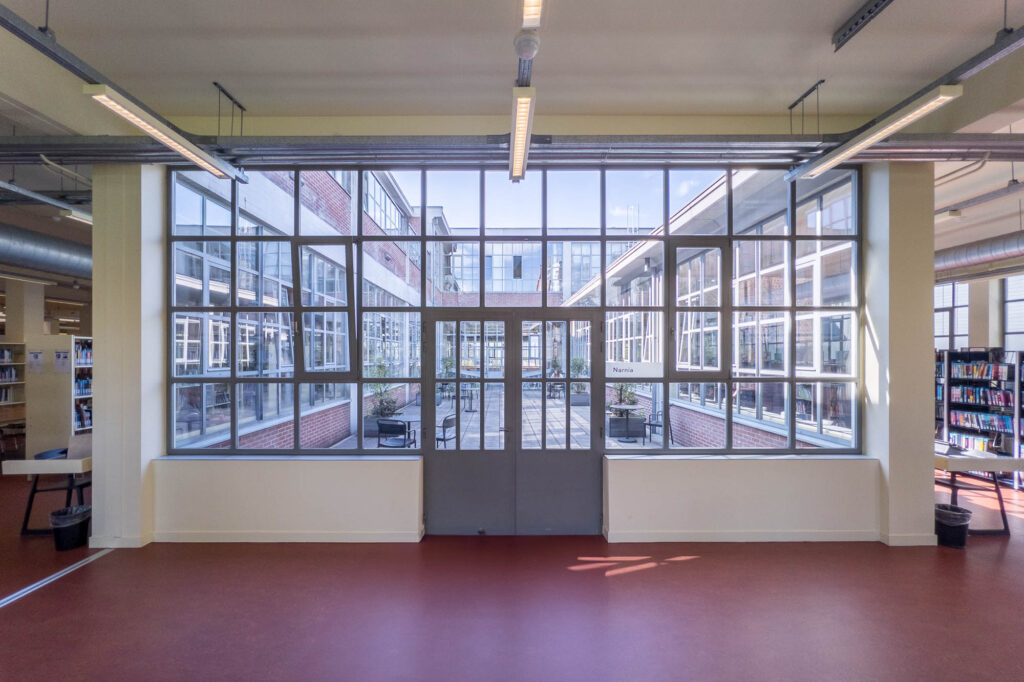 Library interior inside a converted industrial space with large windows. At the centre of the image is a window and door leading to a sunny courtyard with cafe furtniture. Bookshelves are visible on the sides.