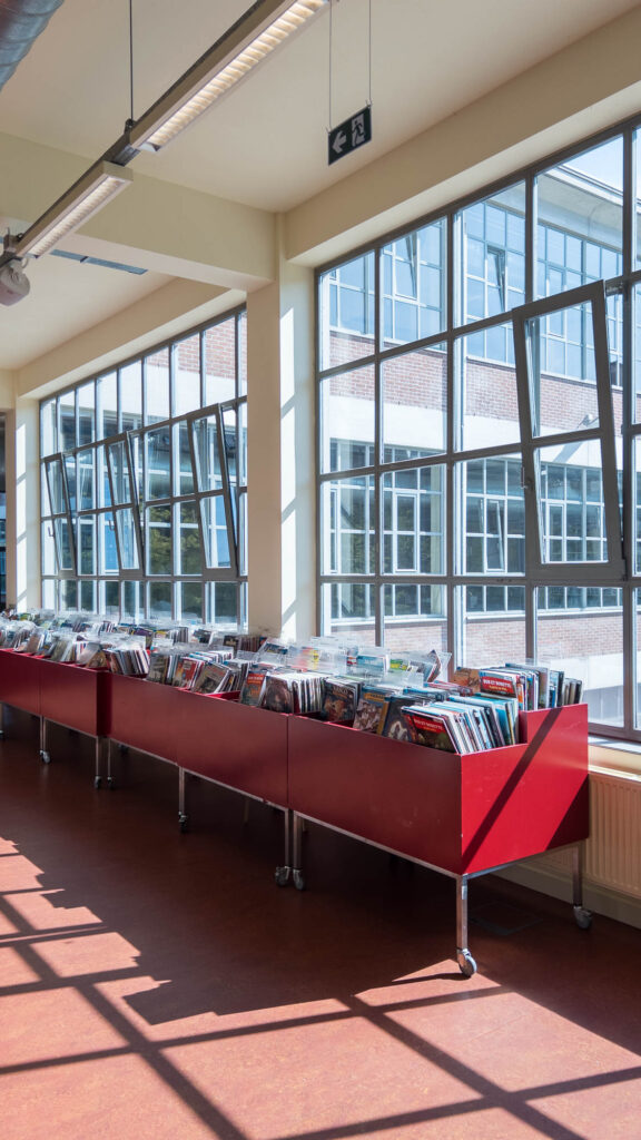 Library interior inside a converted industrial space with large windows. A row of low boxes on casters filled with comic books is in front of a window.