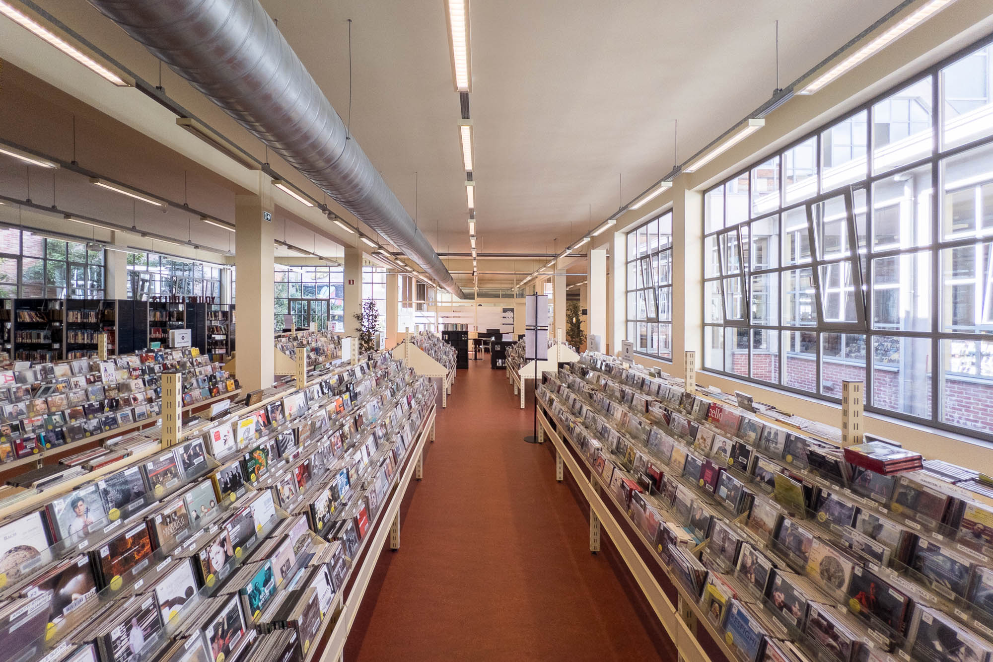 Library interior inside a converted industrial space with large windows. The image looks down an aisle with rows of bookshelves filled with CDs or DVDs. Large windows line the right side, letting in natural light. The ceiling has industrial-style ventilation.