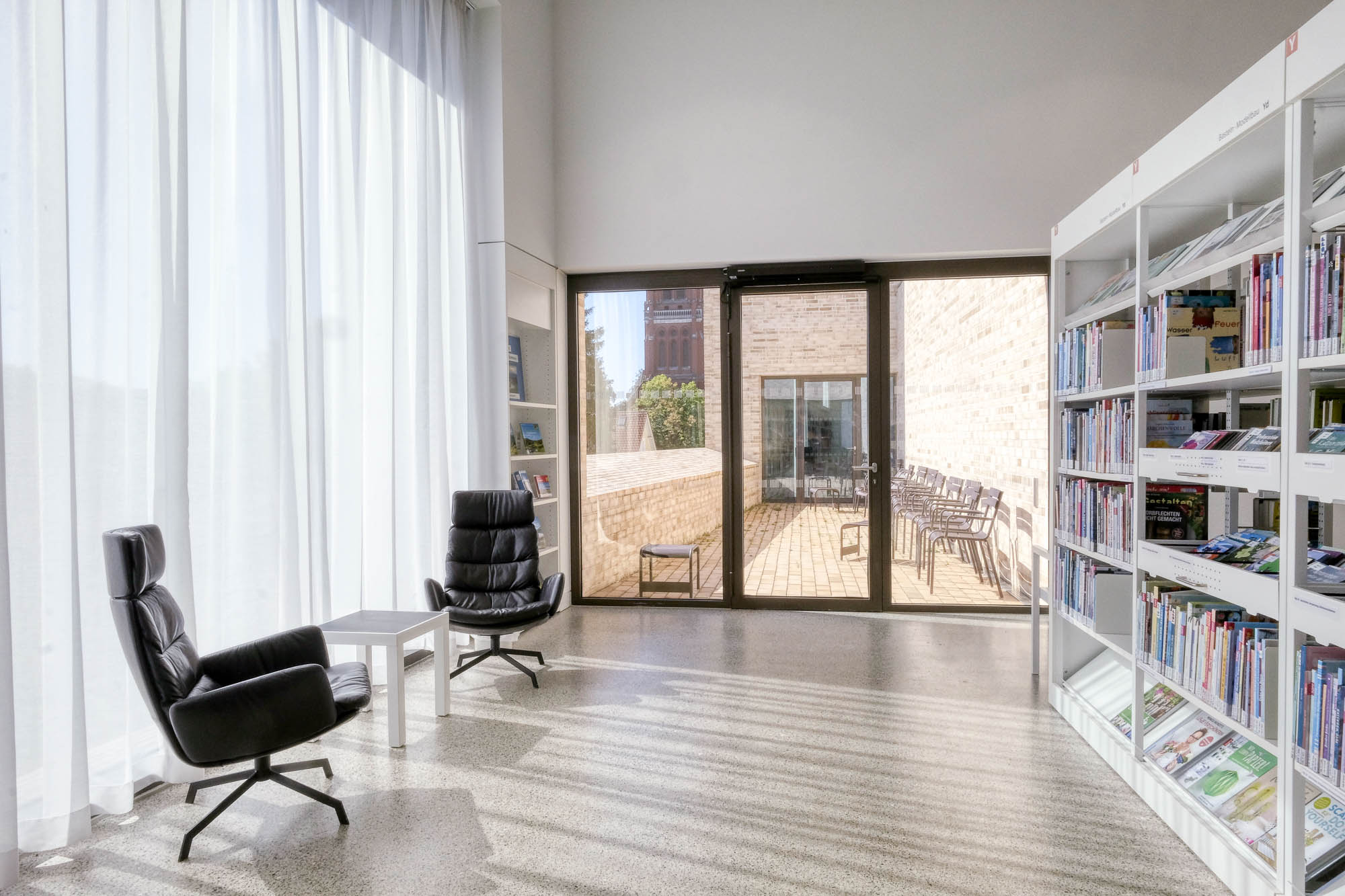 A sunlit library corner with two black leather chairs and a small table near tall windows. Bookshelves line the right wall, and a glass door leads to an outdoor patio with chairs. Light filters softly through sheer curtains.