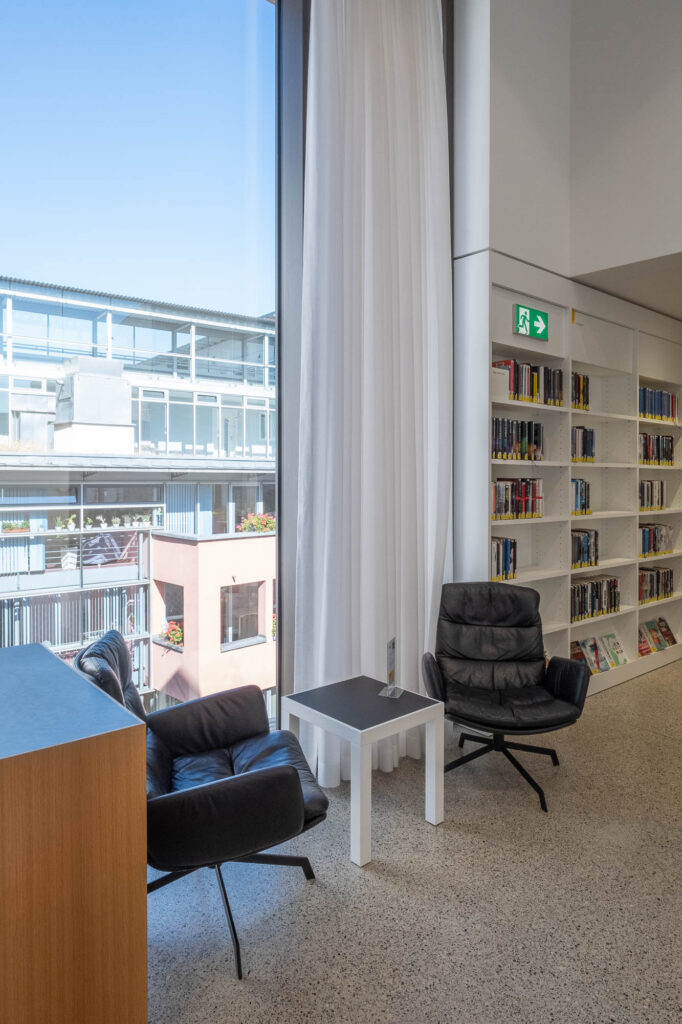 Interior of a contemporary library with a large window and a white bookshelf. Comfy seats and a small coffee table stand before the window. Neighbouring housing blocks can be seen through the window.