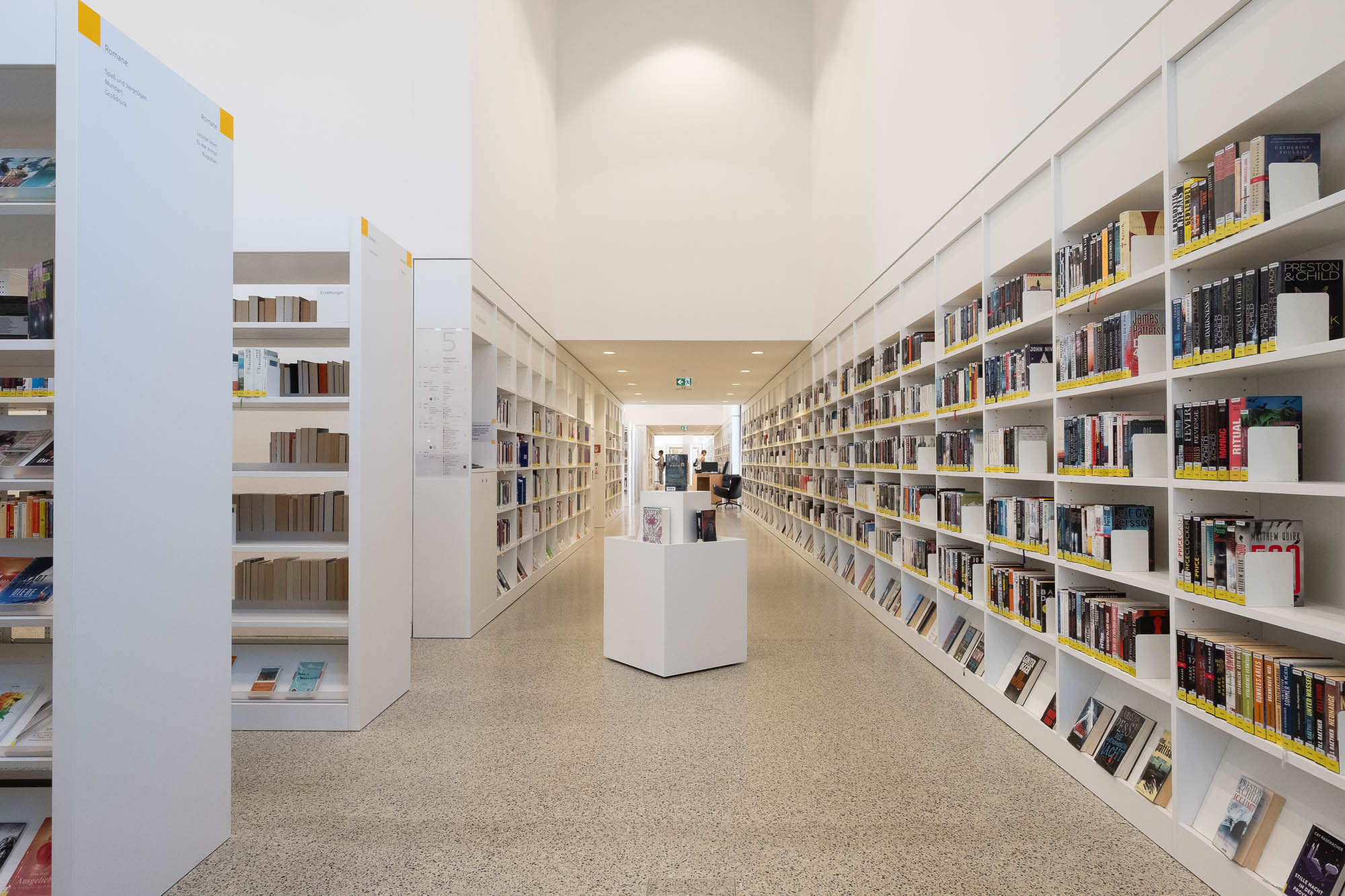 Interior of a contemporary library with bookshelves lining the walls. There is a corridor in the front, with patrons browsing books at the back. A square book display element stands in the foreground.