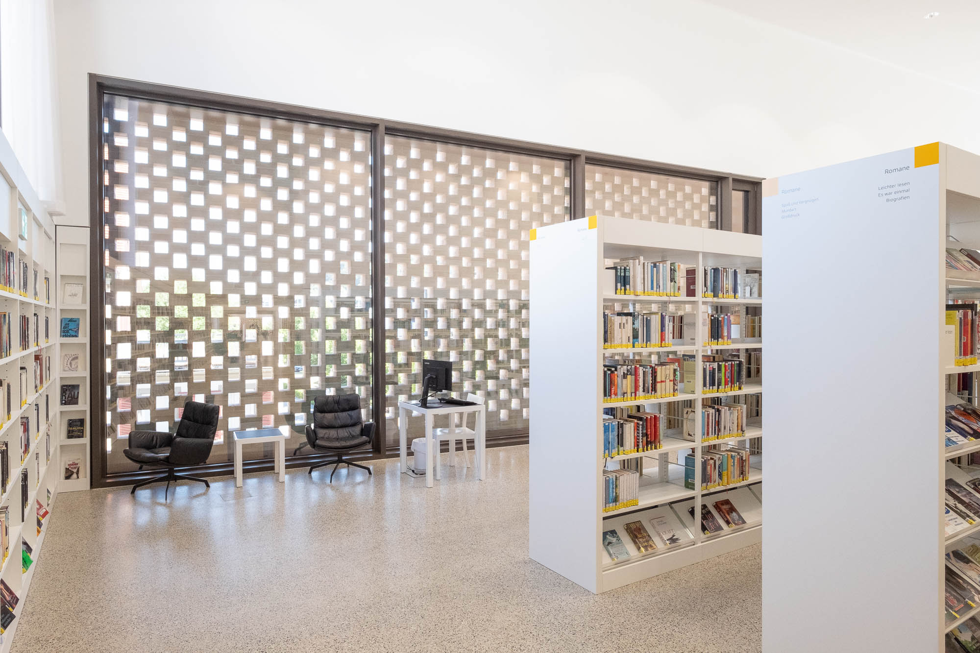 A contemporary library interior with bookshelves holding books. Two black chairs are positioned by a small table with a computer near a large window. In front of the window is a brick lattice letting in only partial light.
