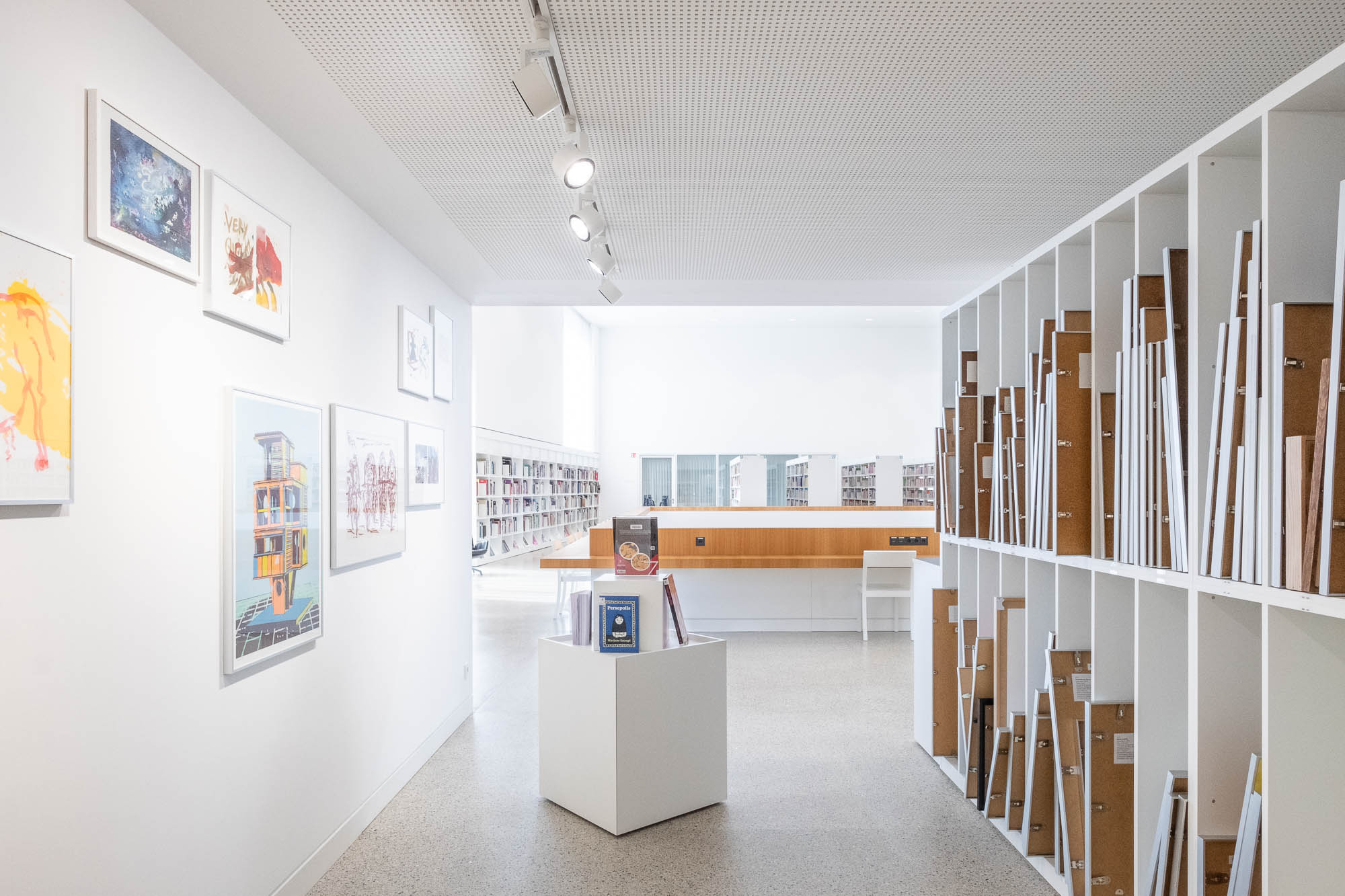 Interior of an art lending section in a contemporary library. There is a white wall displaying framed artwork on the left. Shelving on the right holds large canvases. There is a central pedestal with books. Ceiling lights illuminate the space. Bookshelves are visible in the background.
