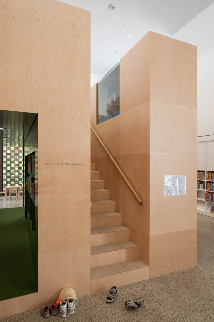 Geometric wooden play structure with a staircase leading to the top. Several pairs of shoes are on the floor at the base of the stairs. A partially visible green-carpeted area with bookshelves appears to the left. A sign advises removing shoes before entering.