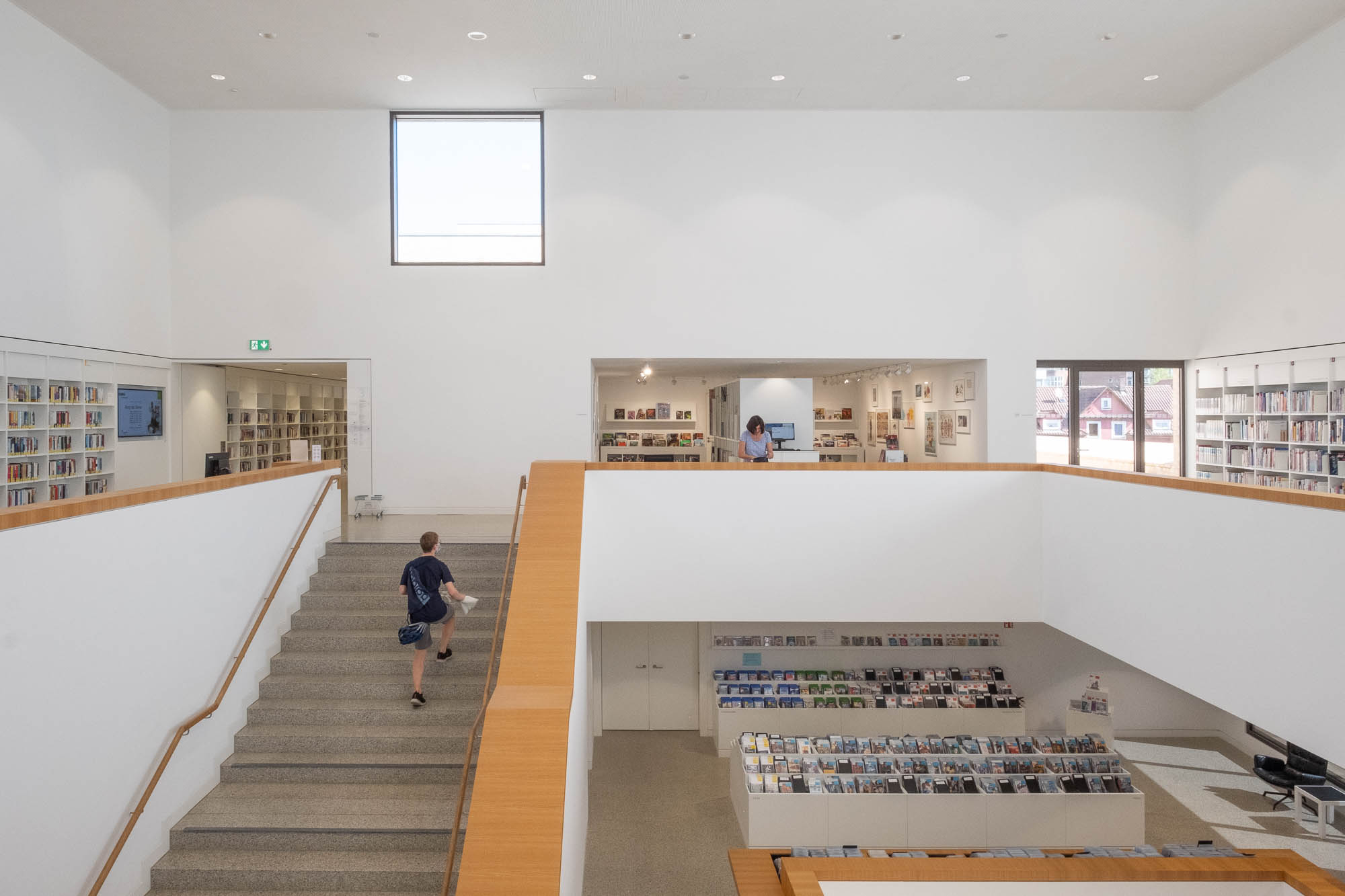 Interior of a contemporary library with white walls. A person walks up a staircase on the left, while another stands by a railing on the upper level. The lower level has boxes of CDs and DVDs. Light comes from a square window high up the wall.