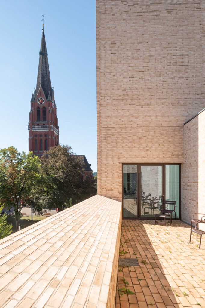 A brick terrace with chairs and a small table, leading to a modern building with glass doors. In the background, a red brick church with a tall steeple is visible under a clear blue sky.