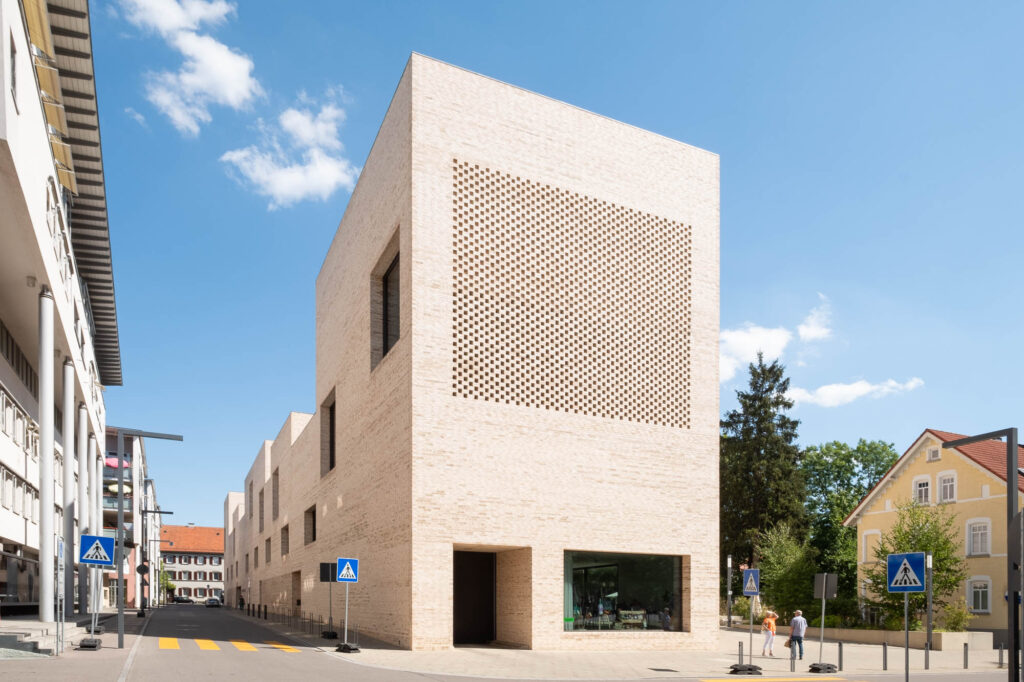 Exterior of a contemporary building with a light brick facade on a street corner with crosswalks. The structure features a perforated design on the upper section. Adjacent buildings and some pedestrians are visible, along with a clear blue sky.