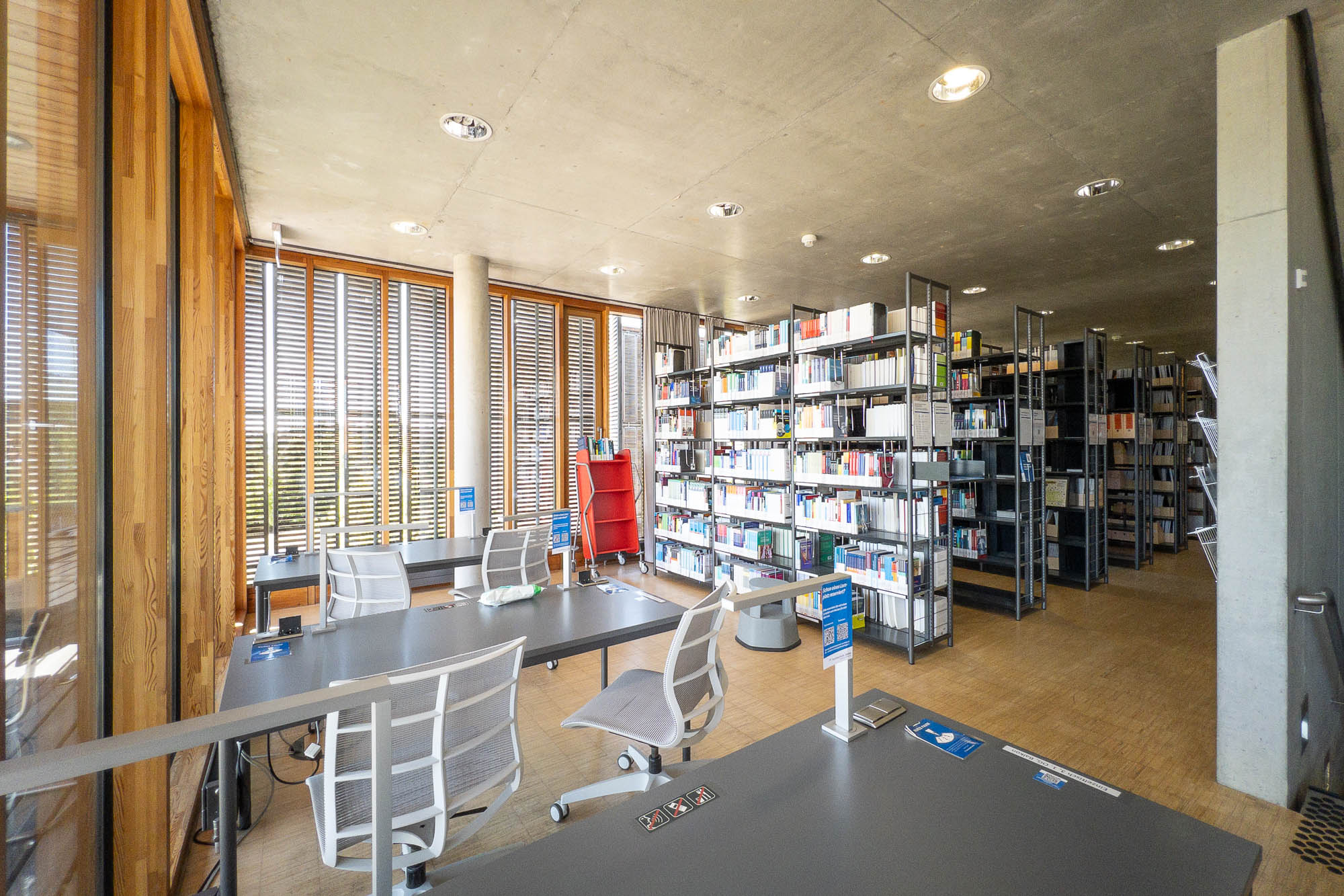 Interior of a contemporary library with concrete walls and wooden floors. There are metal bookshelves at the back and desks with chairs at the front. Windows are shielded from the sun by horizontal wooden blinds.