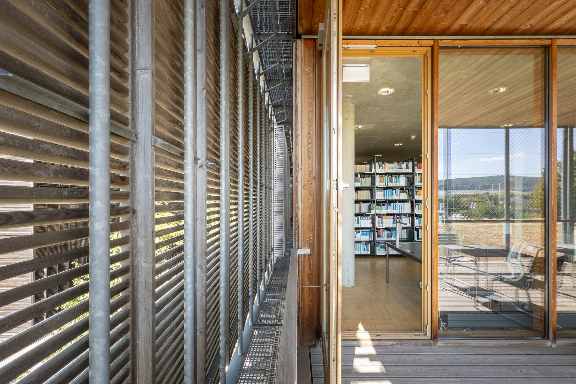 Detail of a contemporary library, showing a gap between the building and wooden blinds on a metal structure. A door is open to the right, and books are visible inside the library.