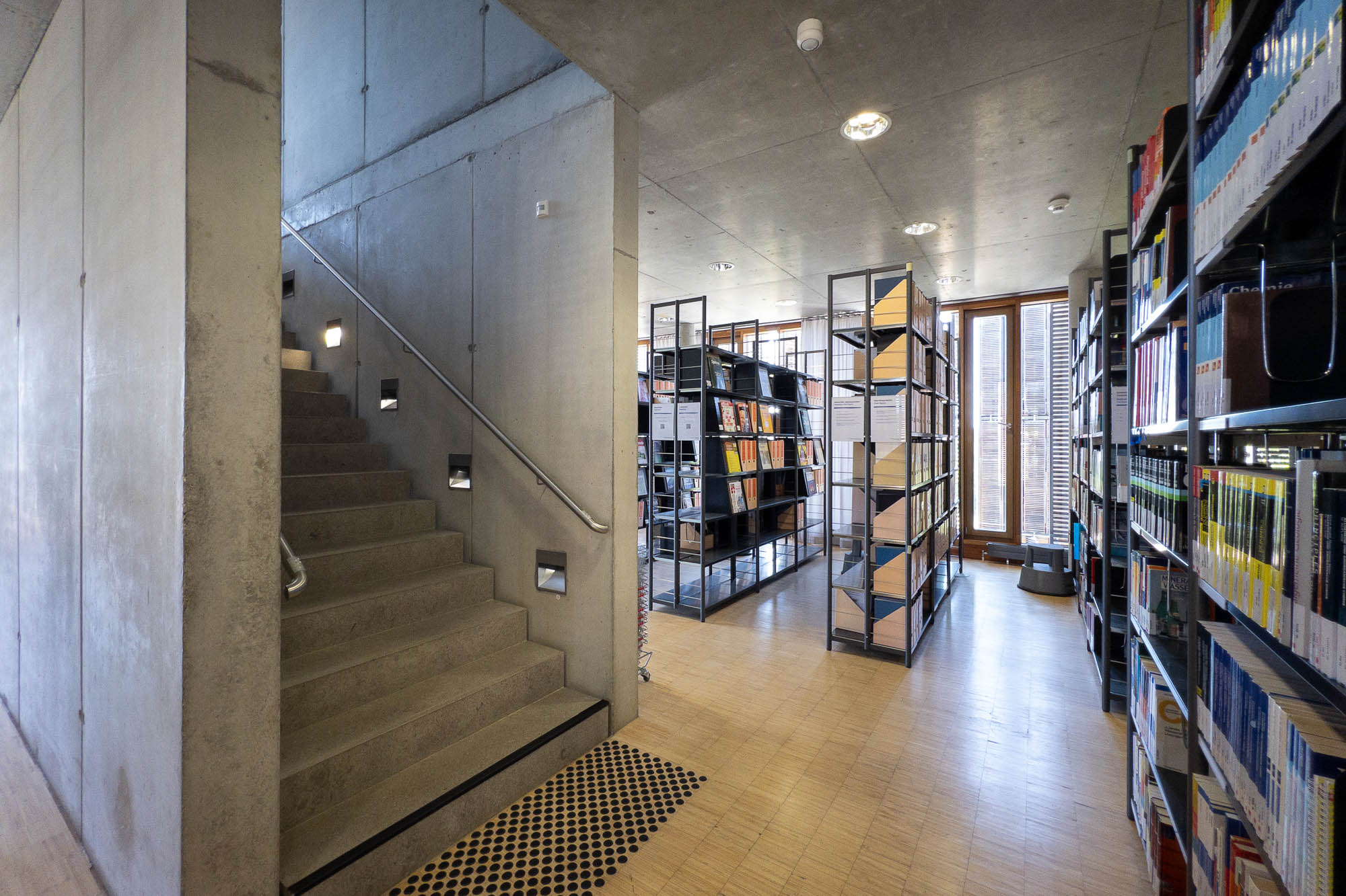 Interior of a contemporary library with concrete walls and wooden floors. A staircase leads to an upper level.