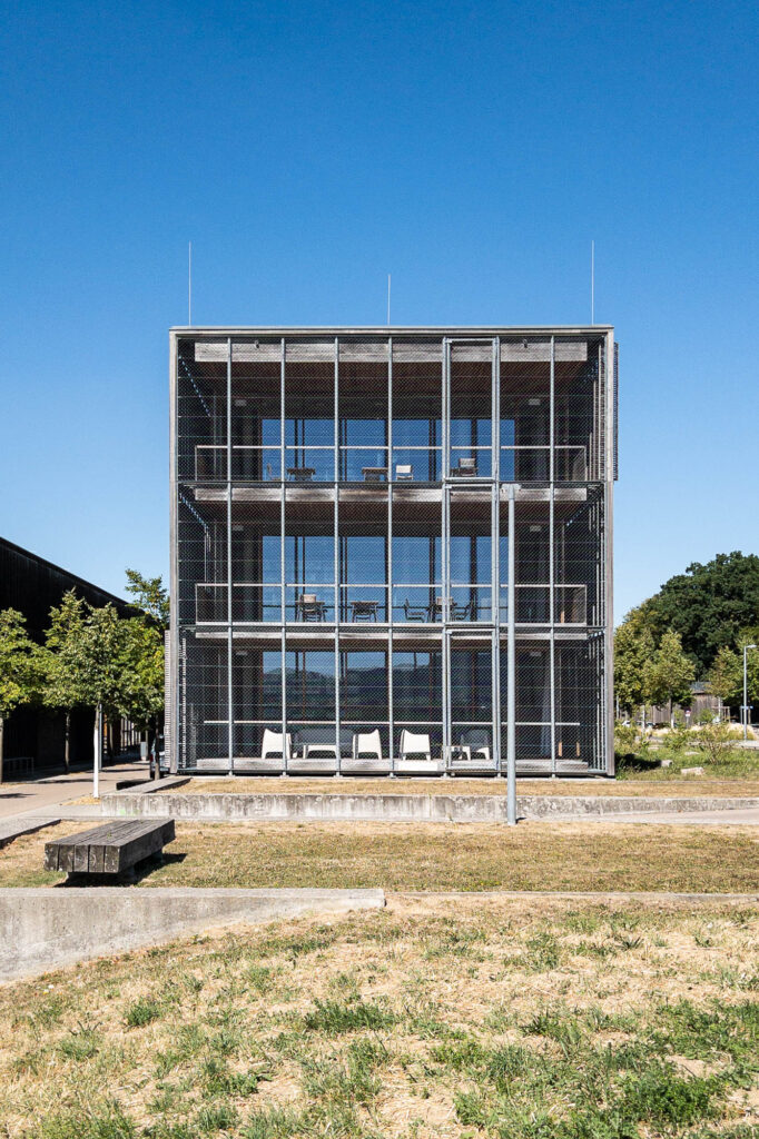 A modern three-story building with a metal grid facade and wooden walls, featuring large windows and balconies on each floor.