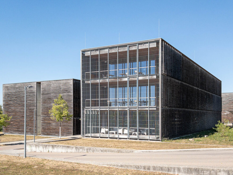 A modern three-story building with a metal grid facade and wooden walls, featuring large windows and balconies on each floor. The structure is surrounded by other similar buildings and shrubs.