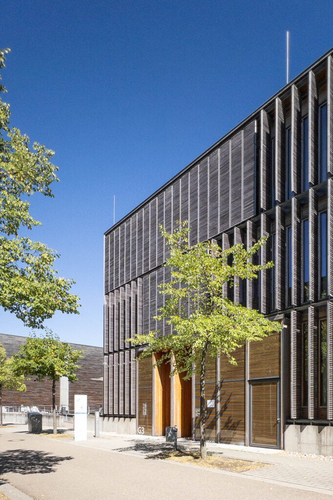 Contemporary building with a grid-like facade and large vertical windows, covered with angled wood lattice blinds. The structure is three stories high, with a bright blue sky in the background. There is a pathway lined with small trees in front.
