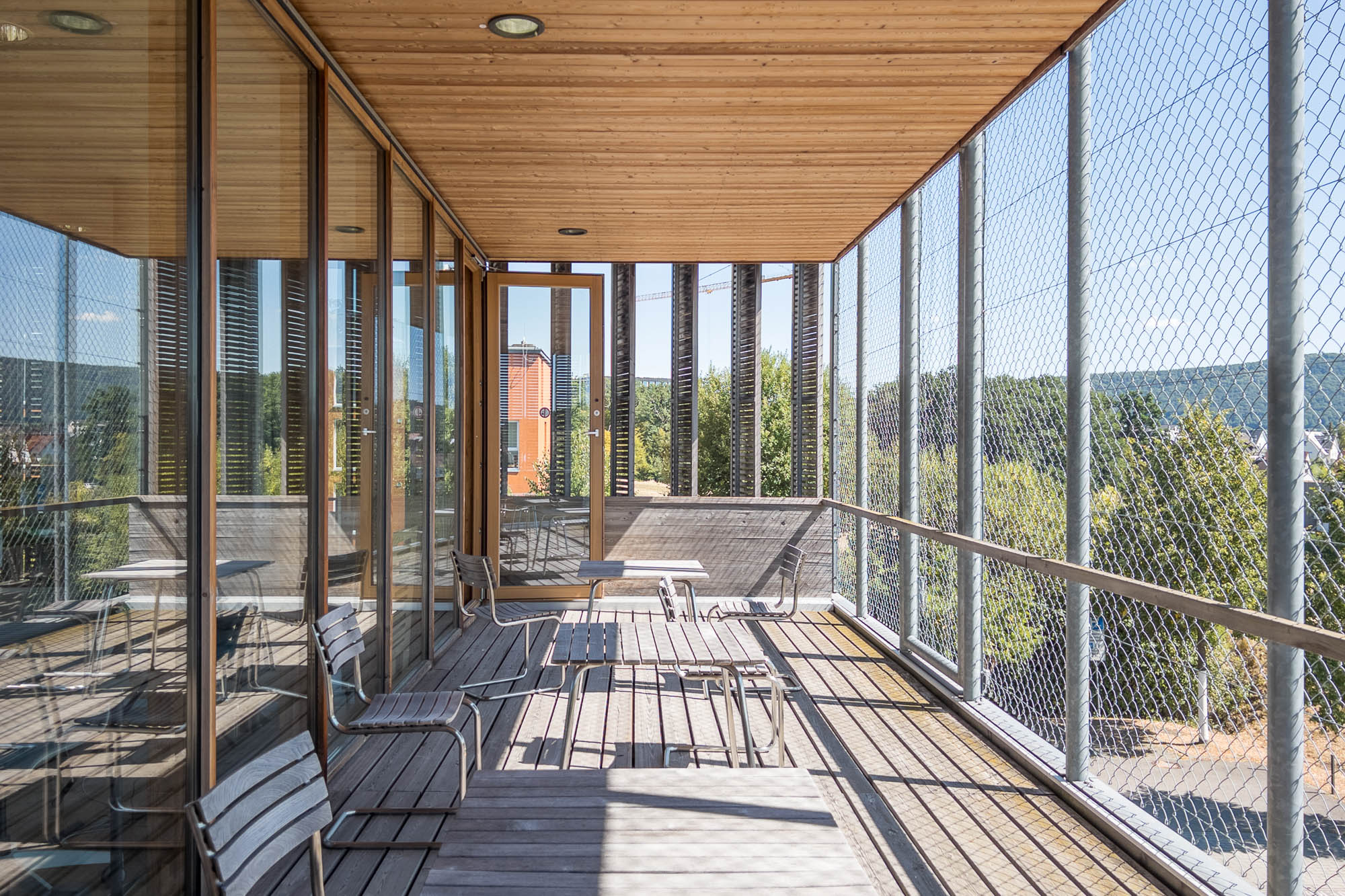 A wooden balcony with glass panels and netted safety rails overlooks a scenic view of greenery and buildings. The space has several metal chairs and tables under a wooden canopy. The sky is clear, and sunlight casts shadows on the floor.