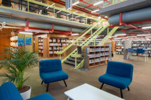 Interior of a 1970s library with blue armchairs, a white table, and a potted plant in the foreground. Shelves filled with books are visible, along with a staircase leading to an upper level. Bright lighting and exposed beams and pipes adorn the ceiling.