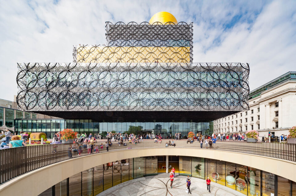 The image shows the modern Library of Birmingham with a gold-topped upper floor and decorative circular metalwork on its facade. People are gathered outside and on the entrance plaza under a partly cloudy sky.